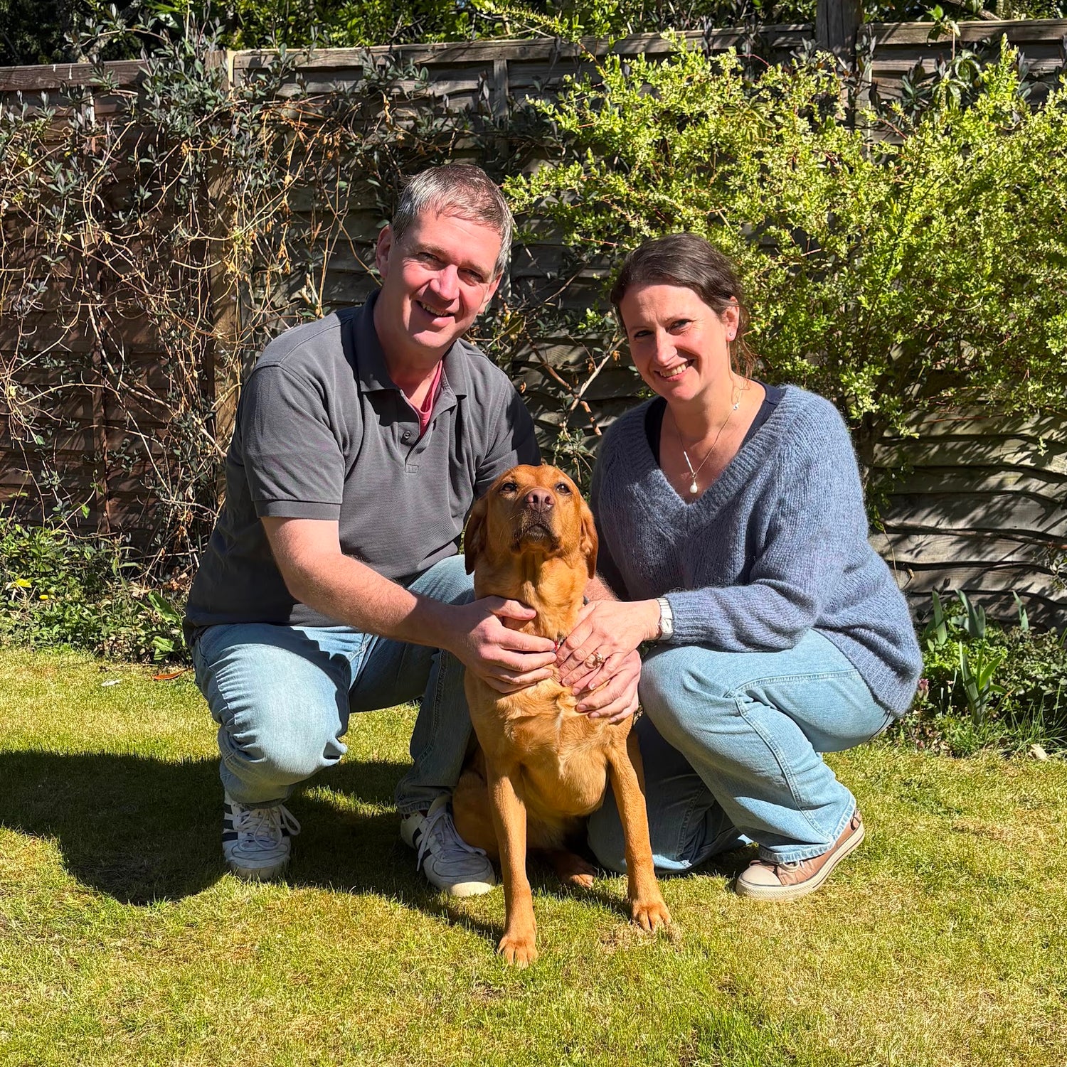 Vets Humphrey and Joanna Grimmett pictured side by side in a garden with their fox red labrador sat in front of them. 